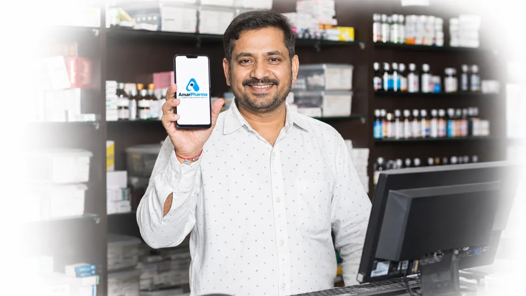 হাতে AmarPharma অ্যাপ খোলা ফোন নিয়ে ফার্মেসির কাউন্টারে একজন ফার্মাসিস্ট